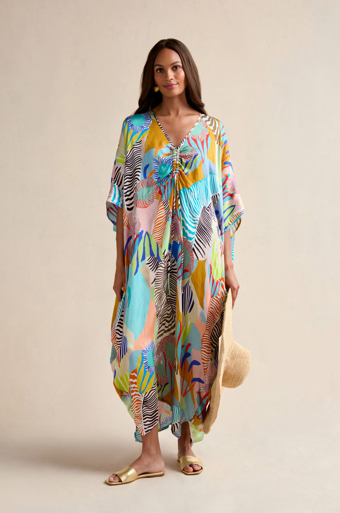 A woman stands against a neutral background wearing the Tropical Fantasy Kaftan with gold sandals, holding a straw hat by her side.