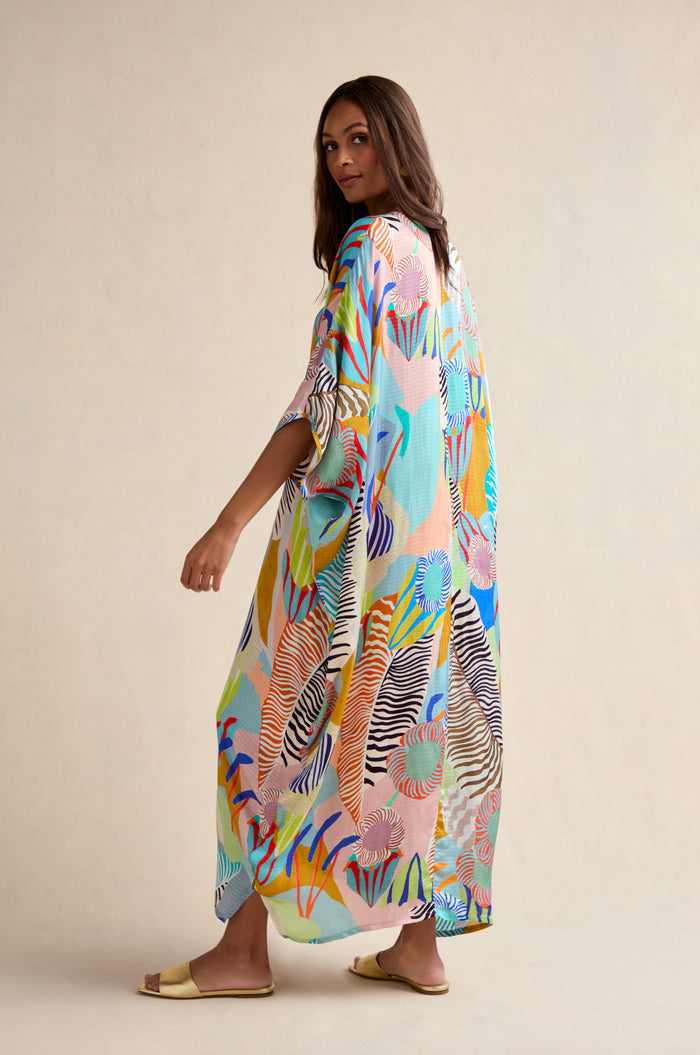 A woman in the Tropical Fantasy Kaftan stands sideways against a neutral background, looking over her shoulder. The oversized maxi dress features colorful patterns, paired with sandals.