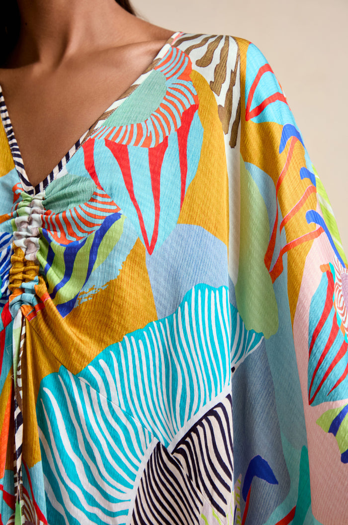A person wearing the Tropical Fantasy Kaftan, featuring bold floral and abstract prints in blue, red, yellow, and green tones with a gathered chest detail.