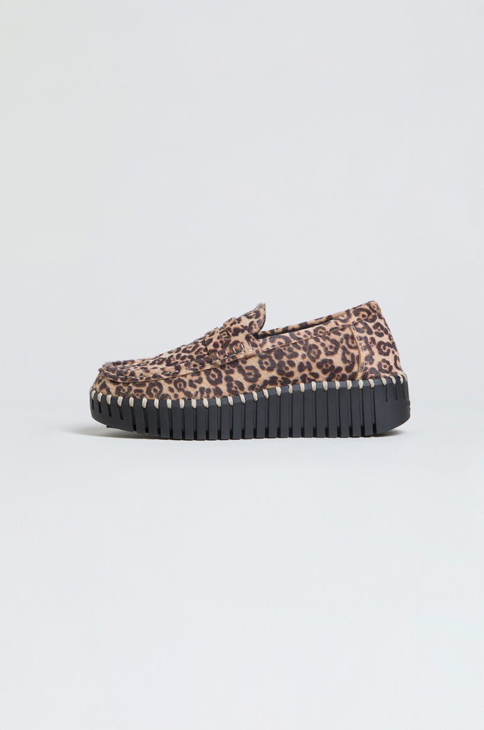 The Tulip Leopard Penny Loafer, featuring a bold leopard print upper and a chunky black ridged wedge sole, is displayed against a plain light background, offering a modern twist on the classic leopard-print loafer.