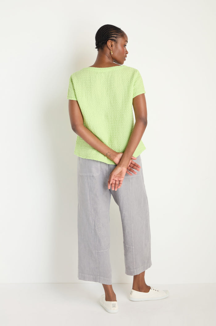 A woman stands against a white wall, facing away, wearing the Waffle Linen Lime V Neck Top, loose gray pants, and white sneakers. Her hands are clasped behind her back.