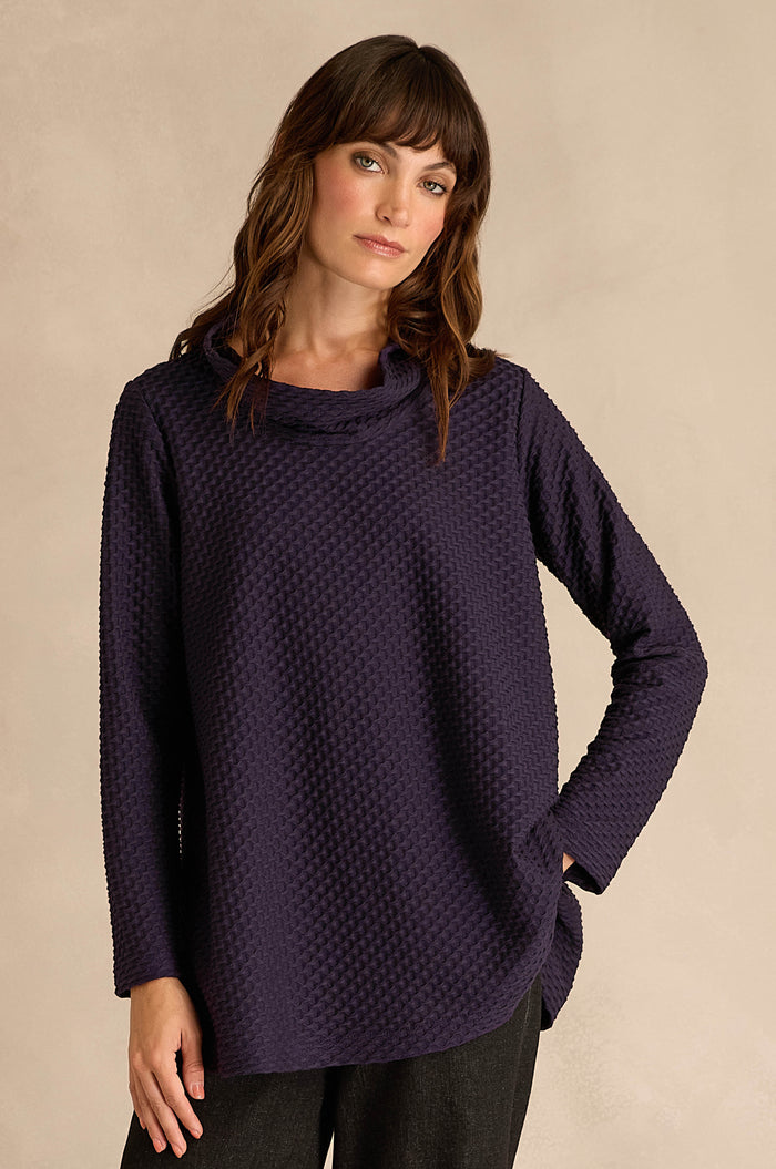 A woman stands against a plain beige background with one hand in her pocket, wearing the Waffle Textured Jersey Damson Cowl Neck Top and dark pants.