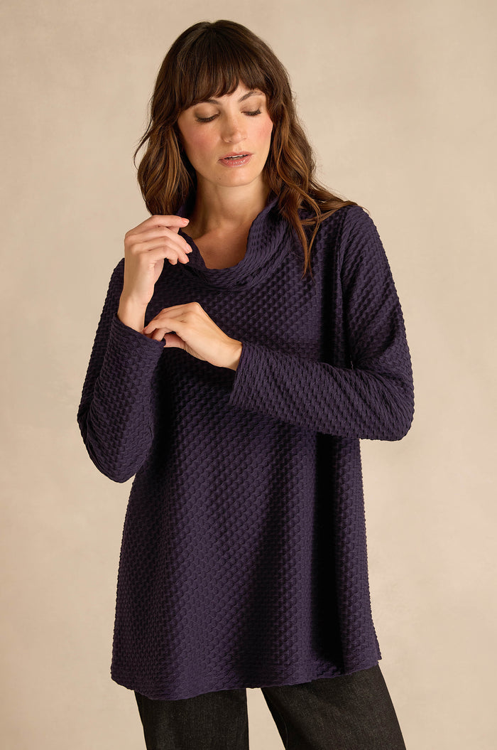 Wearing the Waffle Textured Jersey Damson Cowl Neck Top and black pants, a person stands against a beige background with eyes closed, one hand raised near their face.