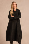 A woman with long blonde hair stands with arms crossed, wearing the Wave Textured Jersey Cowl Neck Dress in black and black leggings, set against a plain beige background.