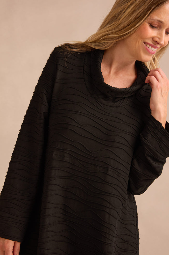 A woman wears the Wave Textured Jersey Cowl Neck Dress in black, featuring long sleeves and a textured finish, as she smiles and looks down against a neutral background.
