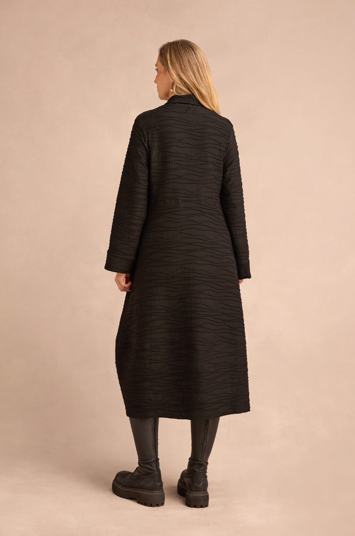 Facing away from the camera, a person wears the Wave Textured Jersey Cowl Neck Dress styled as a coat, paired with black leggings and chunky black shoes against a beige background.