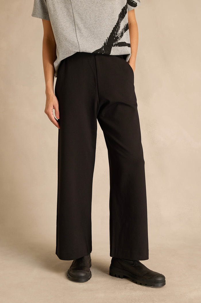 A person stands against a neutral background wearing a grey short-sleeve shirt, the Wide Leg Ponte Trousers in black, and black shoes.