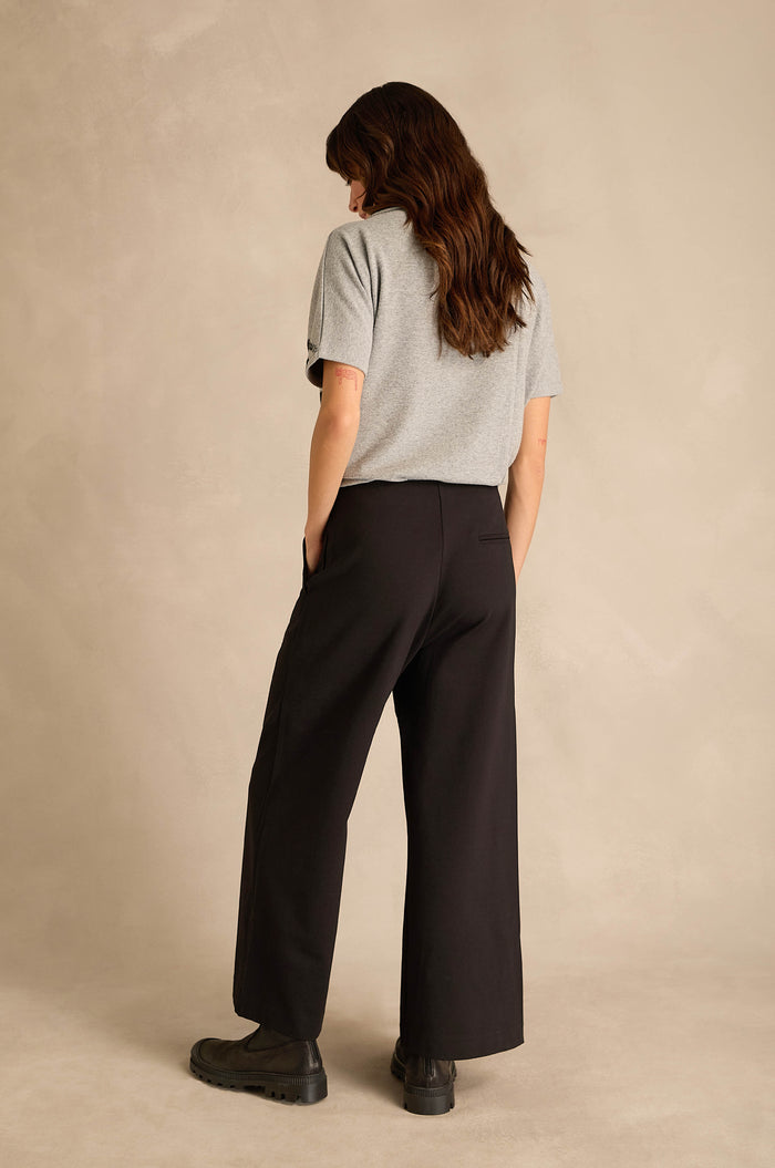 A person stands with their back to the camera, wearing a gray short-sleeve shirt, black shoes, and the Wide Leg Ponte Trousers—black wide leg trousers crafted from stretch ponte jersey—set against a neutral background.