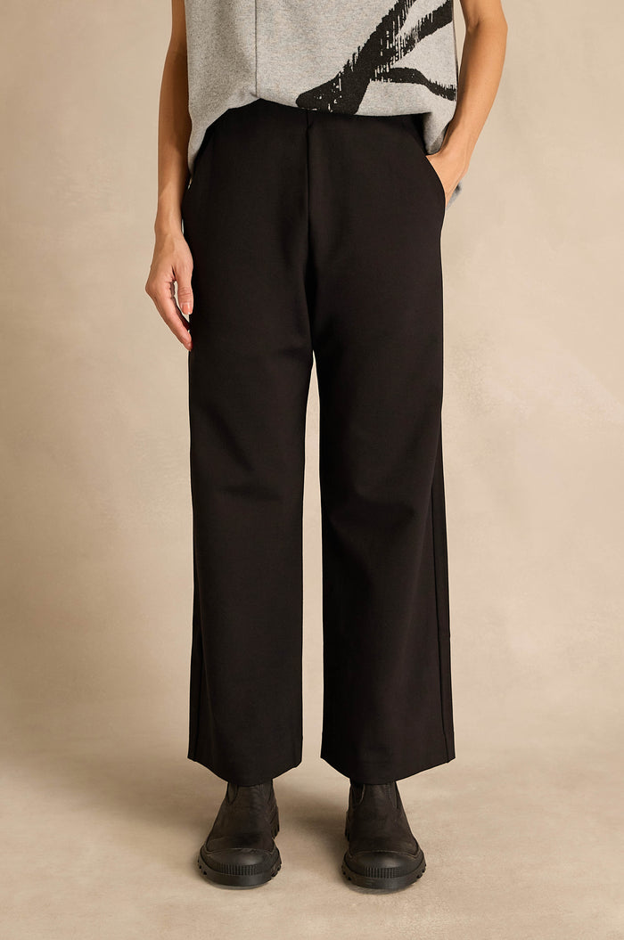 A person stands against a neutral background, wearing a grey top, black Wide Leg Ponte Trousers, and black shoes, with hands in pockets.