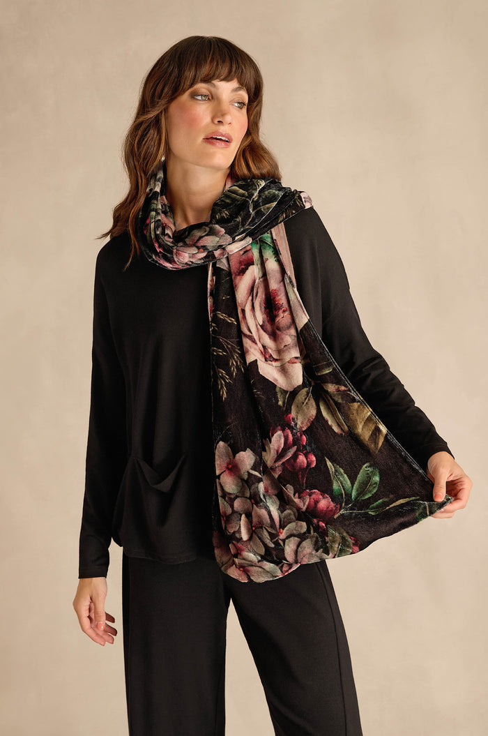 A woman stands against a neutral background, wearing a black long-sleeve top and pants, with the Winter Rose Velvet Scarf draped around her neck and held out to show its elegant rose print.