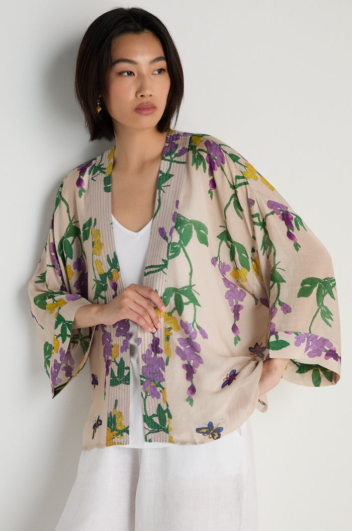 A woman wears the Wisteria Kimono—a light jacket with a purple and green floral print—over a white top and white pants, standing against a plain light-colored wall.
