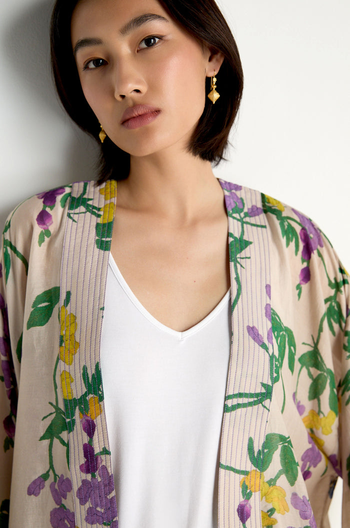 A woman wears the Wisteria Kimono, featuring a tan botanical print with purple and yellow flowers and green leaves, layered over a white V-neck shirt.