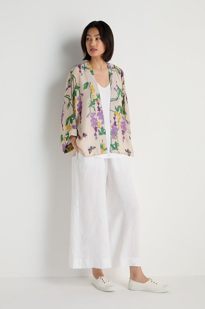 A person stands against a white wall wearing the Wisteria Kimono over a white top and wide-legged white pants, completed with crisp white slip-on shoes.