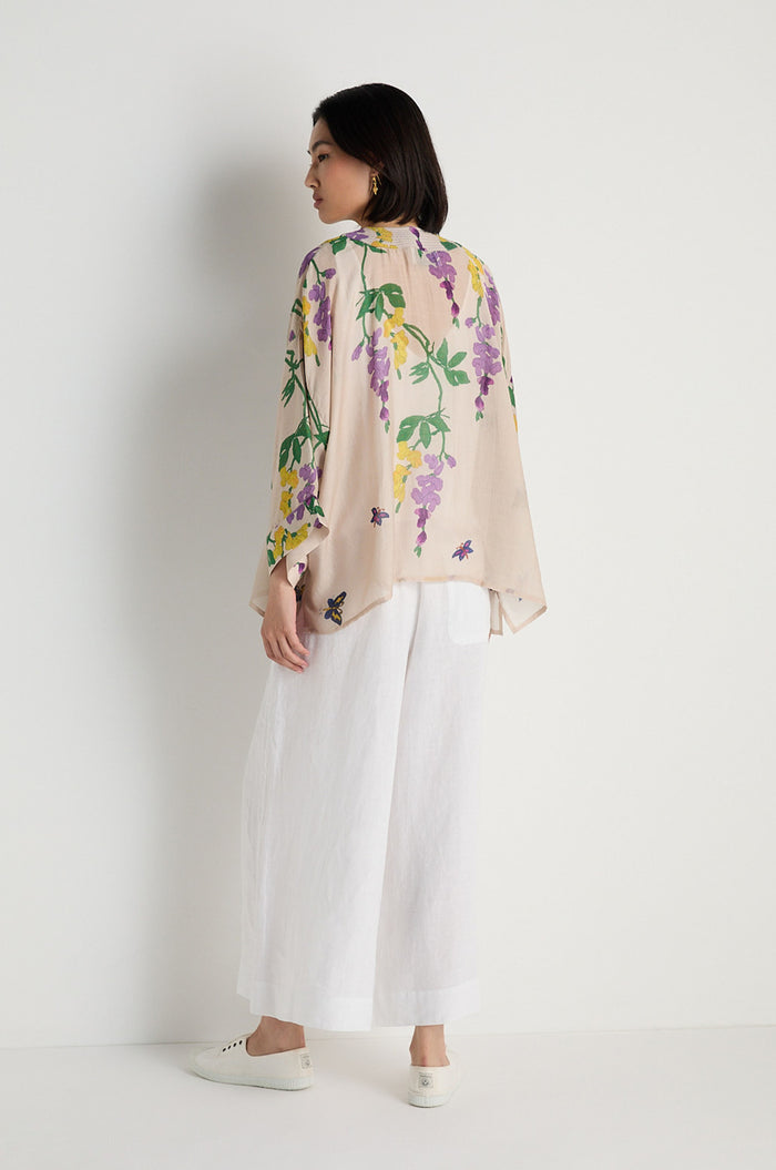 A woman faces partly away from the camera against a white wall, wearing the Wisteria Kimono with a light botanical print over loose white pants and white shoes.