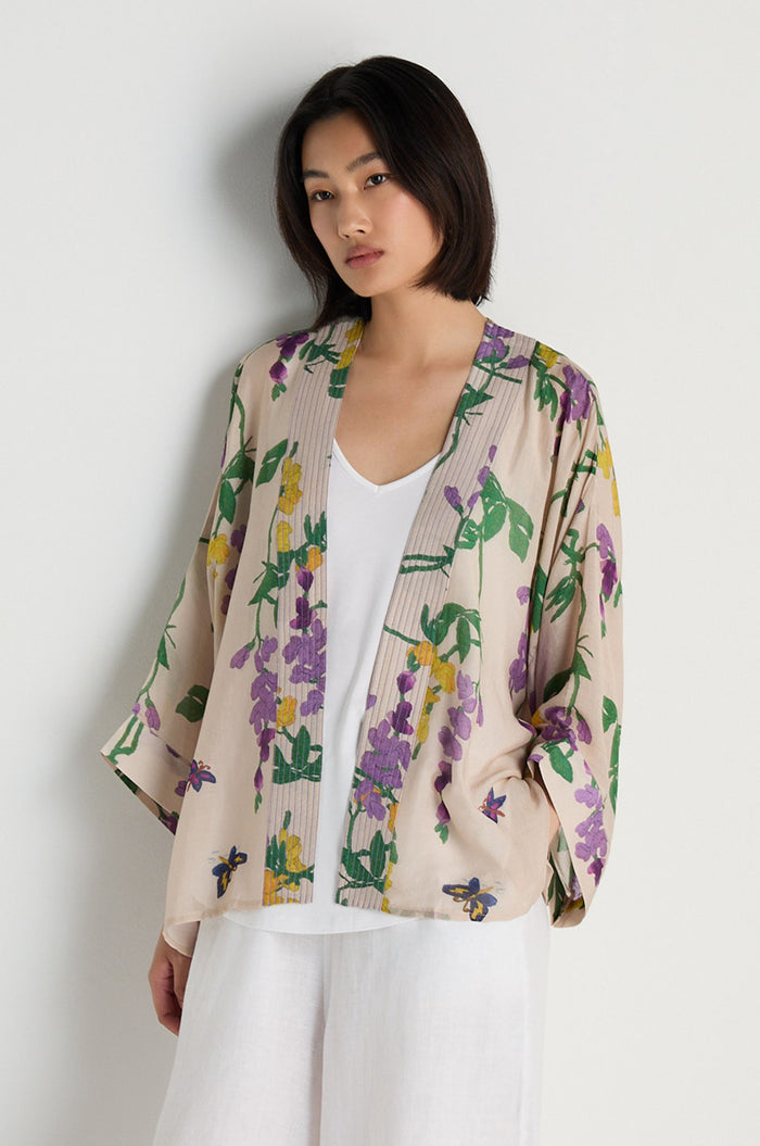 A woman models the Wisteria Kimono, featuring a cream base with purple and yellow floral patterns, layered over a white top and white pants against a plain white background.